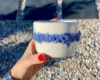 May include: A white ceramic mug with a textured blue band around the middle. The mug is held in a hand with red nail polish. The background is out of focus, with a mix of pebbles and stone.