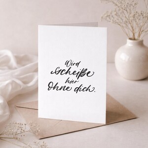 May include: A white greeting card with black calligraphy that reads "Wird Scheiße hier Ohne dich." The card is on a brown envelope, with a white cloth and a vase with dried flowers in the background.