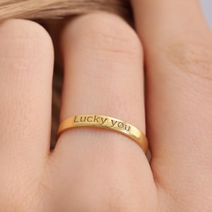 Può includere: Un anello dorato con l'iscrizione "Lucky you" è mostrato su un dito. L'anello è sottile e ha una superficie lucida. Lo sfondo è sfocato, focalizzando l'attenzione sull'anello.
