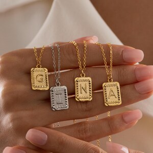 May include: A collection of gold and silver initial pendant necklaces. Each necklace features a rectangular pendant with a letter of the alphabet. The pendants are suspended from delicate gold chains. The necklaces are displayed against a white background.