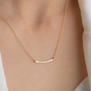 May include: A delicate gold-colored necklace with a curved bar pendant. The pendant has engraved text: "45.0390 N 79.3079 W". The necklace is worn around the neck, and the chain is visible. The background is a person's skin and white clothing.