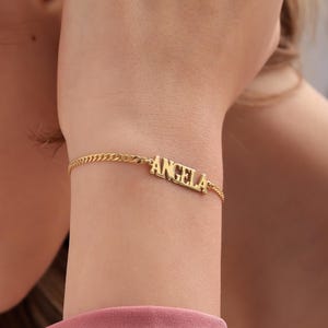 May include: A gold chain bracelet with the name "ANGELA" spelled out in block letters.
