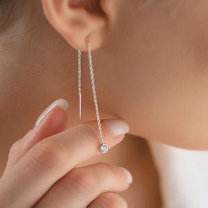 Op de afbeelding: Een close-up van een zilveren threader-oorbel met een kleine, ronde, heldere edelsteen aan het uiteinde. De oorbel hangt aan het oor. De andere oorbel is ook zichtbaar.