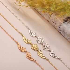 May include: Three delicate chain bracelets in rose gold, gold, and silver. Each bracelet features a different charm: wings, a flower, and a rose. The bracelets are displayed on a light beige surface.