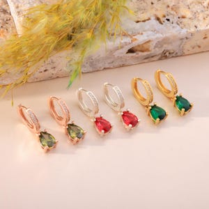 May include: Six pairs of hoop earrings with teardrop-shaped gemstones in various colors: olive green, red, and emerald green. The hoops are gold, silver, and rose gold, with small clear stones. The earrings are displayed on a light pink surface.