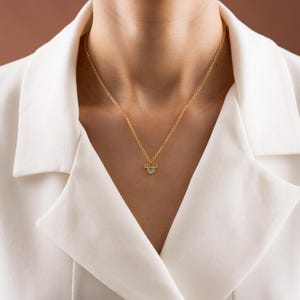 以下が含まれることがあります： マウスの頭の形をしたペンダントが付いたゴールドカラーのネックレス。ペンダントには、3つの透明な丸い宝石が付いています。白いブレザーの上からネックレスを着用しています。