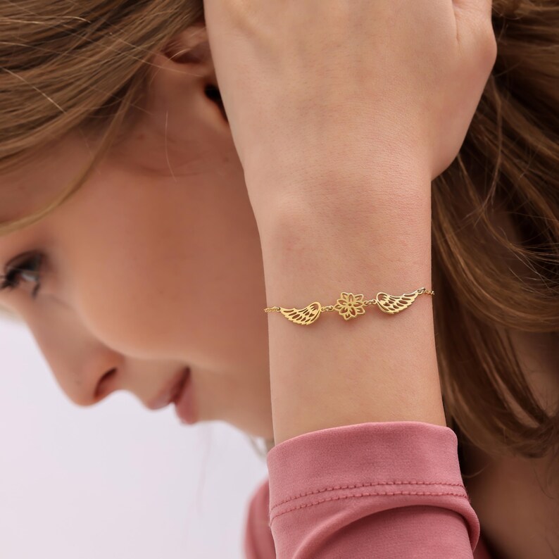 May include: A gold-colored bracelet featuring a floral design in the center, flanked by two wing-shaped charms. The bracelet is delicate and appears to be made of a thin chain. The bracelet is worn on a wrist.