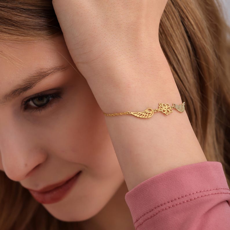 May include: A delicate gold bracelet featuring three charms: a wing, a flower, and a leaf. The bracelet is worn on a wrist, with the charms displayed prominently. The gold chain is thin and elegant, creating a minimalist aesthetic.