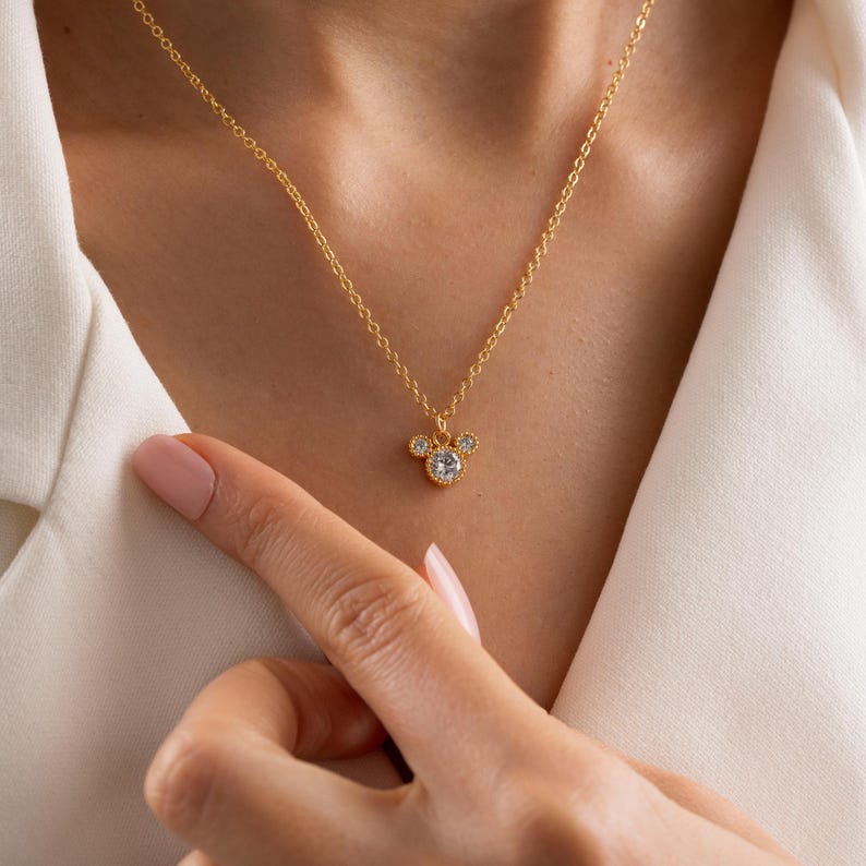 以下が含まれることがあります： 金色のネックレス。ペンダントはネズミの頭の形をしており、中央に大きな透明な石があり、その周りを小さな透明な石が囲んでいます。白いシャツを着た人がネックレスを着用しています。