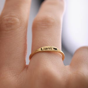 Peut inclure: Une bague dorée gravée du prénom "Liam" et d'un symbole de cœur. La bague est portée au doigt, mettant en valeur le détail fin de l'inscription personnalisée. Le design de la bague est simple et élégant.