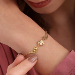 May include: A gold-colored bracelet featuring a delicate design of wings and a floral element. The bracelet is worn on a wrist, showcasing its intricate details and elegant style. The jewelry is a beautiful accessory.