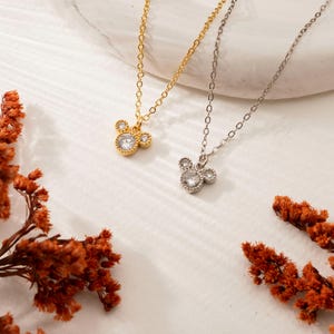 以下が含まれることがあります： マウスの頭の形をしたペンダントチャームが付いた2つのネックレス。1つは金色、もう1つは銀色です。マウスヘッドペンダントには、透明でキラキラ光る石がちりばめられています。ネックレスは、乾燥したオレンジ色の花が添えられた白い表面に展示されています。