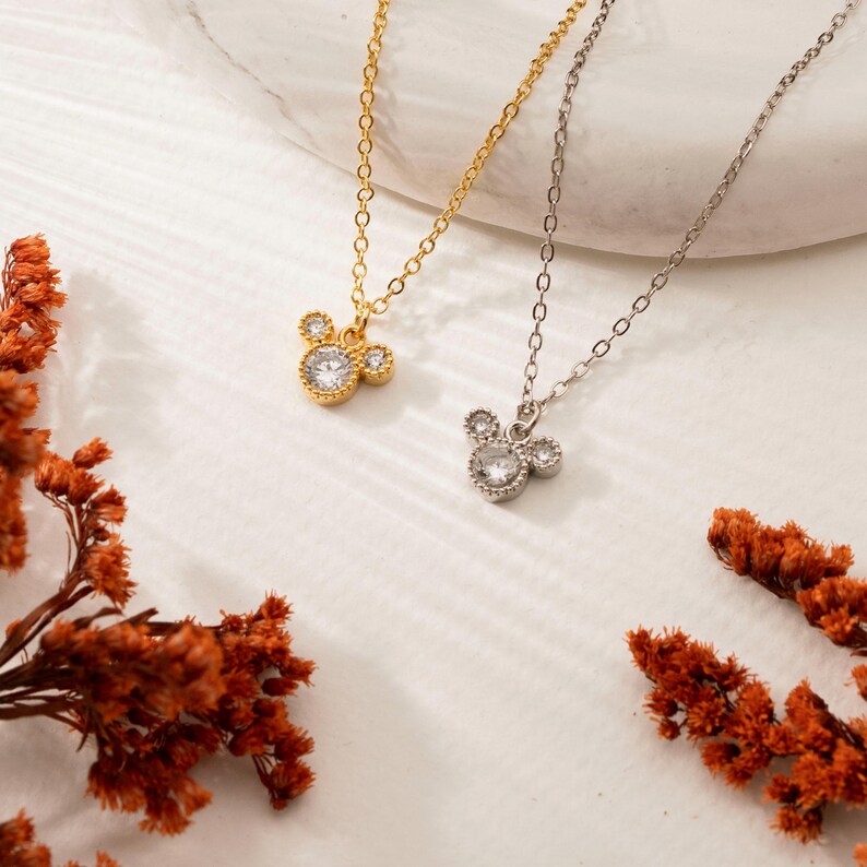 以下が含まれることがあります： マウスの頭の形をしたペンダントチャームが付いた2つのネックレス。1つは金色、もう1つは銀色です。マウスヘッドペンダントには、透明でキラキラ光る石がちりばめられています。ネックレスは、乾燥したオレンジ色の花が添えられた白い表面に展示されています。