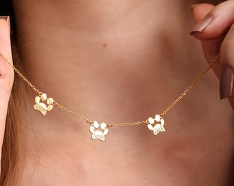 May include: A gold-colored necklace featuring three paw print charms, each with a name engraved. The names are Liam, Noah, and Jack. The necklace is displayed against a blurred background.