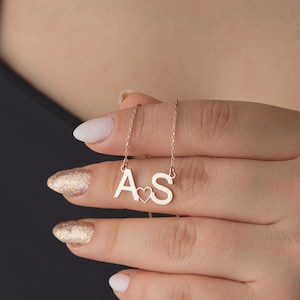 May include: A rose gold necklace featuring the letters "A" and "S" with a heart symbol between them. The pendant hangs from a delicate chain. The necklace is displayed on a person's hand.