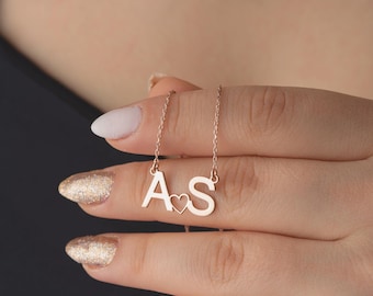 May include: A rose gold necklace featuring the letters "A" and "S" with a heart symbol between them. The pendant hangs from a delicate chain. The necklace is displayed on a person's hand.