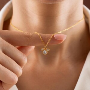 以下が含まれることがあります： 金のネックレス。ペンダントはマウスの頭の形をしており、中央に大きな丸い透明な宝石があり、その周りを小さな透明な宝石が囲んでいます。ネックレスは人の指で持ち上げられています。