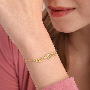 May include: A gold-colored bracelet featuring a delicate design. The bracelet has a chain with three decorative elements: a pair of wings, a flower, and another pair of wings. The bracelet is worn on a wrist.