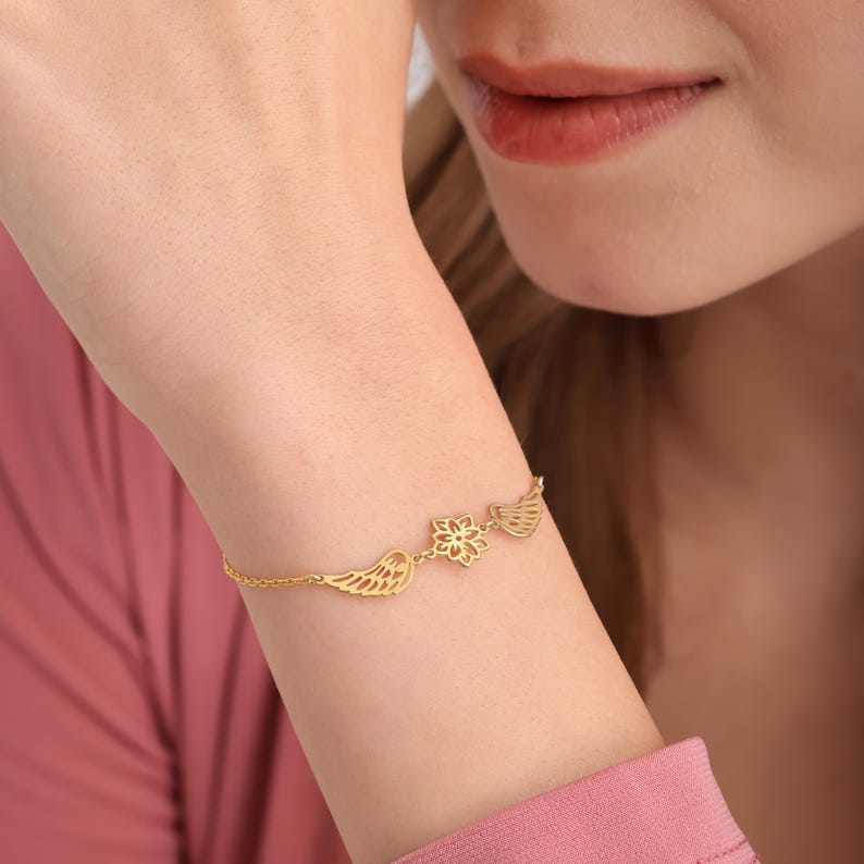 May include: A gold-colored bracelet featuring a delicate design. The bracelet has a chain with three decorative elements: a pair of wings, a flower, and another pair of wings. The bracelet is worn on a wrist.