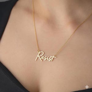 May include: A gold-colored necklace with the word "Resist" in a cursive font. The necklace is worn around a person's neck, with the chain and pendant clearly visible against the skin and a black top.