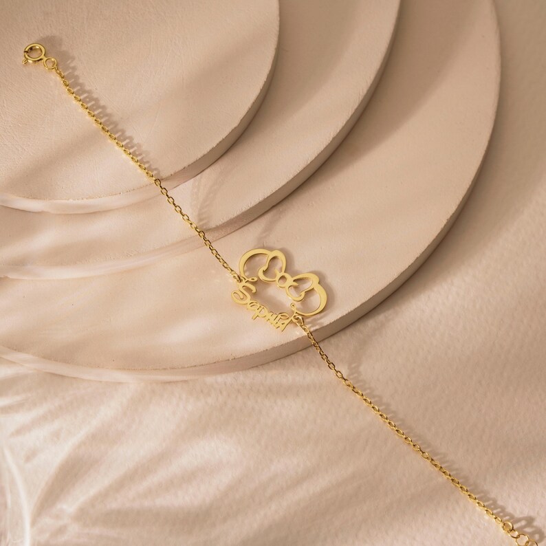 May include: A delicate gold bracelet with a bow-shaped charm and the name "Sophia" in cursive. The fine chain bracelet is displayed on a neutral surface. The bracelet is a stylish piece of jewellery.