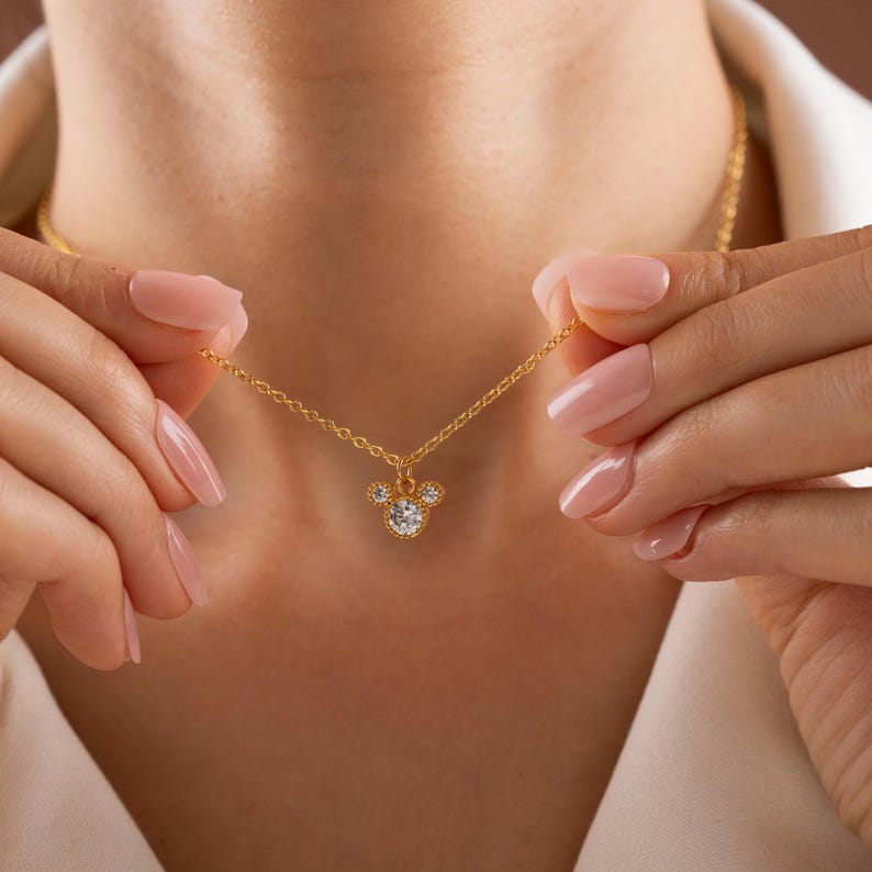 以下が含まれることがあります： 金色のネックレスで、ミッキーマウスの頭の形をしたペンダントが付いています。ペンダントの中央には、大きくて丸い透明な宝石があり、その周りを小さな透明な宝石が囲んでいます。ネックレスは人が持っています。