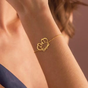 May include: A gold-coloured bracelet with a bow design and the name "Sophia" in cursive. The fine chain bracelet is worn on a wrist, displaying the personalised jewellery.
