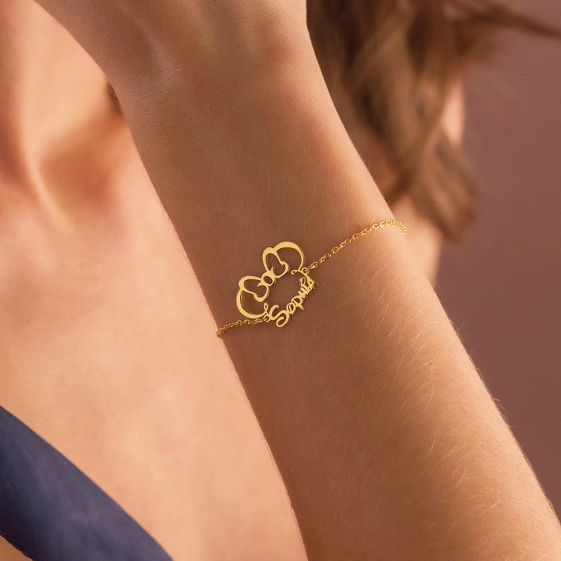 May include: A gold-coloured bracelet with a bow design and the name "Sophia" in cursive. The fine chain bracelet is worn on a wrist, displaying the personalised jewellery.