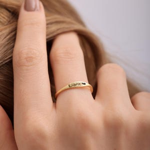 Könnte beinhalten: Ein goldfarbener Ring mit dem eingravierten Namen "Liam" und einem kleinen Herzsymbol. Der Ring wird an einem Finger getragen und zeigt sein schlichtes, elegantes Design. Ein zarter Schmuck, ideal als personalisiertes Geschenk.