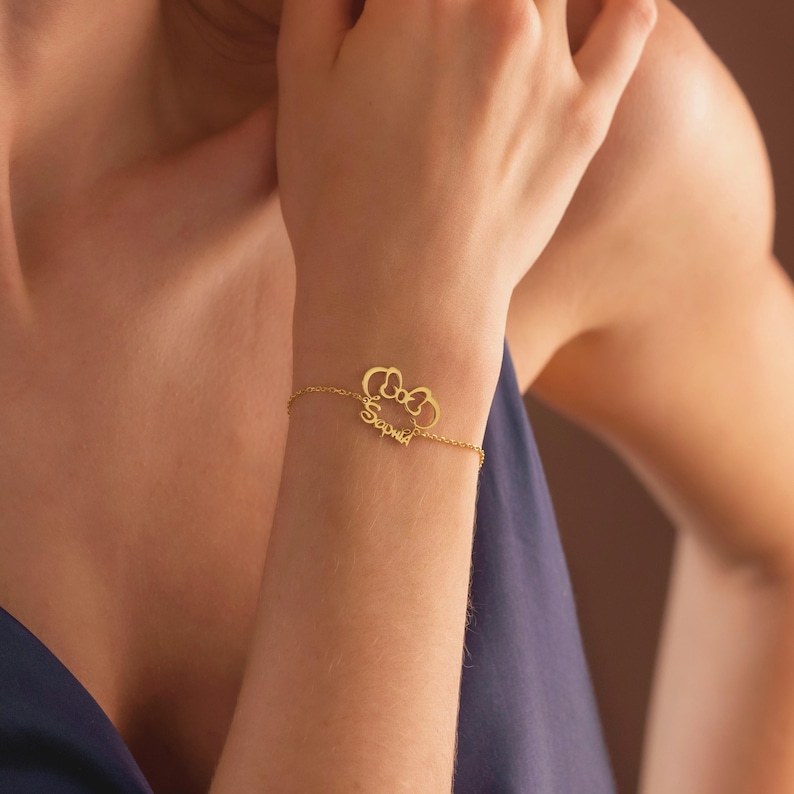 May include: A gold bracelet featuring a bow-shaped pendant and the name "Sophia" in cursive. The fine chain bracelet is displayed on a wrist, highlighting the personalised jewellery item.