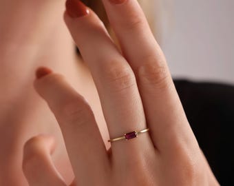 May include: A delicate gold ring featuring a rectangular, deep red gemstone. The ring's band is thin and minimalist, showcasing the vibrant color of the stone. The ring is worn on a finger, highlighting its elegant design.