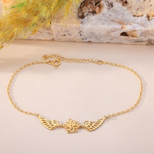 May include: A delicate gold-colored anklet featuring a floral design in the center, flanked by two stylized wing motifs. The anklet is adjustable and appears to be made of a fine chain. The background is a soft, neutral color, highlighting the jewelry.