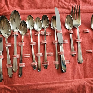 May include: A collection of silver-colored flatware, including knives, spoons, and forks, arranged on a red fabric. The utensils are secured with pink and white woven bands. The set includes various sizes of spoons and knives.