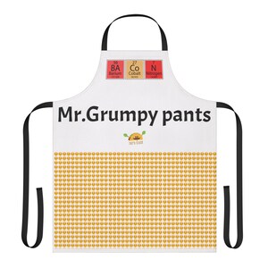 May include: White apron with black straps, featuring the text "Mr. Grumpy Pants" and a cartoon taco with a mustache. The bottom of the apron has a pattern of yellow taco emojis.