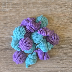 May include: A collection of small, textured seashell-shaped buttons in alternating shades of light blue and purple. Each piece features a detailed, ridged surface pattern and a spiral or fan-like shell structure, arranged in a loose, overlapping cluster.