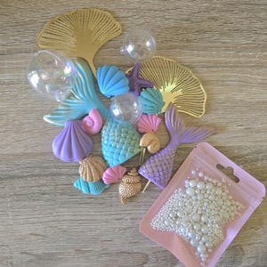 May include: A collection of craft supplies including two gold ginkgo leaf cutouts, two mermaid tails in blue and purple, various plastic seashells in pastel colours, three clear spherical ornaments, and a pink pouch filled with white pearl beads.