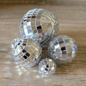 May include: Five silver disco balls of different sizes. Each ball is covered in small, reflective mirrored squares, creating a sparkling effect. The balls are arranged on a wooden surface, reflecting light.