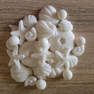 May include: A collection of small, white, matte-finish decorative items shaped like various sea life, including starfish, seashells, spiral conch shells, sand dollars, and smooth spheres, arranged in a cluster on a flat surface.