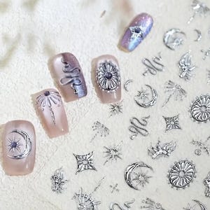 Puede incluir: Primer plano de arte de uñas con diseños celestiales y abstractos plateados. Las uñas están pintadas en un color rosa claro e iridiscente, con adornos plateados que incluyen lunas, soles, estrellas y patrones florales. El tema general es místico y elegante.
