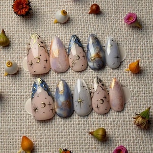 Pode incluir: Um conjunto de dez unhas artificiais com tema celestial. As unhas apresentam desenhos de luas, estrelas e detalhes dourados em uma variedade de cores, incluindo rosa, azul e branco. As unhas estão dispostas sobre uma superfície texturizada com pequenas decorações florais.