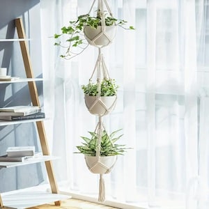 Puede incluir: Colgador de plantas de macramé de tres niveles con macetas de cerámica blancas llenas de vegetación. El colgador está suspendido frente a una cortina blanca transparente, creando una estética luminosa y aireada. El diseño de macramé añade un toque bohemio.