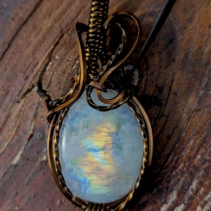 May include: An oval moonstone pendant featuring a translucent white gemstone with iridescent blue and yellow flashes, encased in a dark, oxidized copper wire-wrapped setting with a coiled bail at the top for hanging on a necklace chain.
