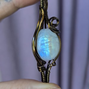 May include: A pendant featuring a smooth, oval-shaped moonstone with a blue iridescent sheen, encased in a dark, oxidized copper wire-wrapped frame. The metalwork consists of intricate, swirling wire coils that secure the stone in place.