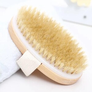 Può includere: Spazzola a secco ovale con setole naturali. Presenta un manico in legno chiaro, un bordo in plastica bianca e un cinturino in tessuto intrecciato. Le setole sono di colore marrone chiaro e la spazzola è progettata per la cura del corpo.