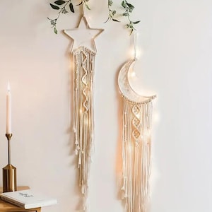 Op de afbeelding: Crèmekleurige macramé wandkleden in de vorm van een ster en een halve maan met lichtslingers. De ster- en maanvormen zijn gemaakt met een geweven ontwerp en lange, vloeiende kwasten. Decoratieve groene planten hangen boven de wandkunst.