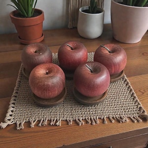 Puede incluir: Cinco manzanas rojas con tallos marrones se asientan sobre pequeños posavasos de madera, dispuestos sobre un mantel individual tejido. Las manzanas son de color rojo intenso y la escena se desarrolla sobre una mesa de madera. Plantas decorativas en macetas de terracota y blancas en el fondo.