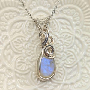 May include: A silver wire-wrapped pendant necklace featuring a teardrop-shaped moonstone with a blue iridescent sheen. The metal wire is intricately coiled into decorative swirls above the stone, suspended from a delicate silver chain.