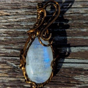May include: A pendant featuring an oval-shaped, translucent moonstone with a blue iridescent sheen, encased in a decorative, dark bronze-colored metal wire-wrapped frame that forms a loop at the top for hanging on a chain or cord.