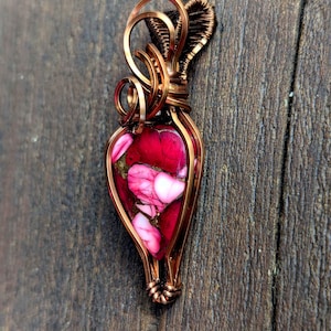 Könnte beinhalten: Handgefertigter Anhänger mit Kupferdrahtrahmen. Der Anhänger zeigt einen tropfenförmigen Stein mit einer lebendigen Mischung aus roten und rosa Farbtönen. Die Drahtarbeit umfasst dekorative Schlaufen und aufwendige Wicklungen.