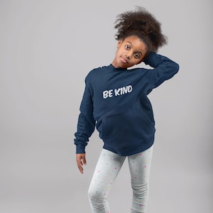 May include: A navy blue hoodie with the words "BE KIND" in white, paired with gray leggings patterned with small hearts. The outfit is completed with pink high-top sneakers.